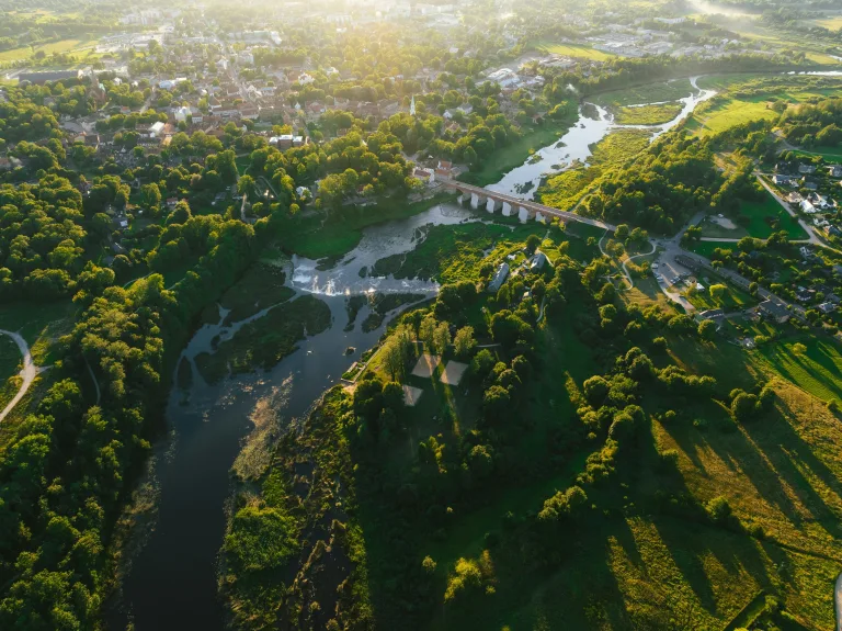 Légi felvétel egy zöldellő, folyóparti kisvárosról naplementekor, középpontban egy híddal és egy kisebb vízeséssel, ami a vonzó lakókörnyezetet és a városfejlesztést szimbolizálja.
