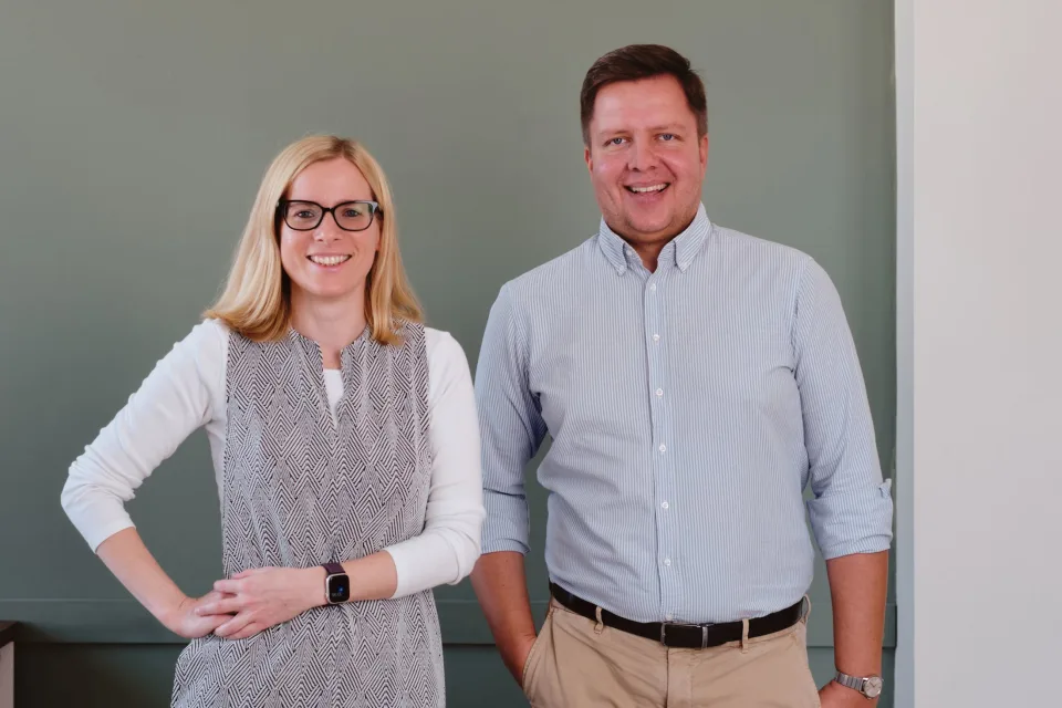 Mosolygó férfi és női ügyvéd, az Illés és Szabó Ügyvédi Társulás vezetői, egy szürke fal előtt állnak.